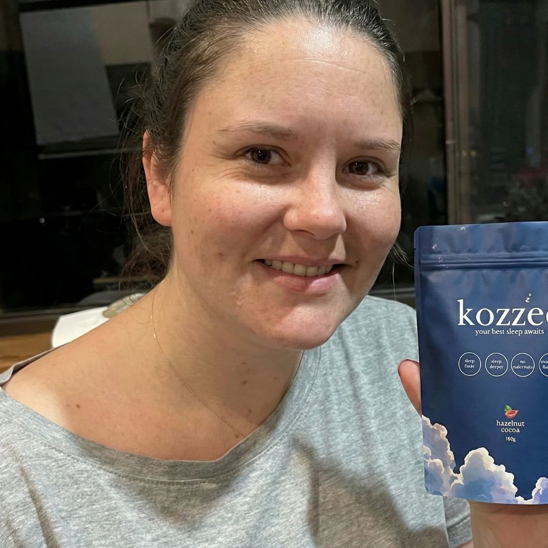 A smiling woman in a grey t-shirt holds up a blue pouch of Kozzee hazelnut cocoa.