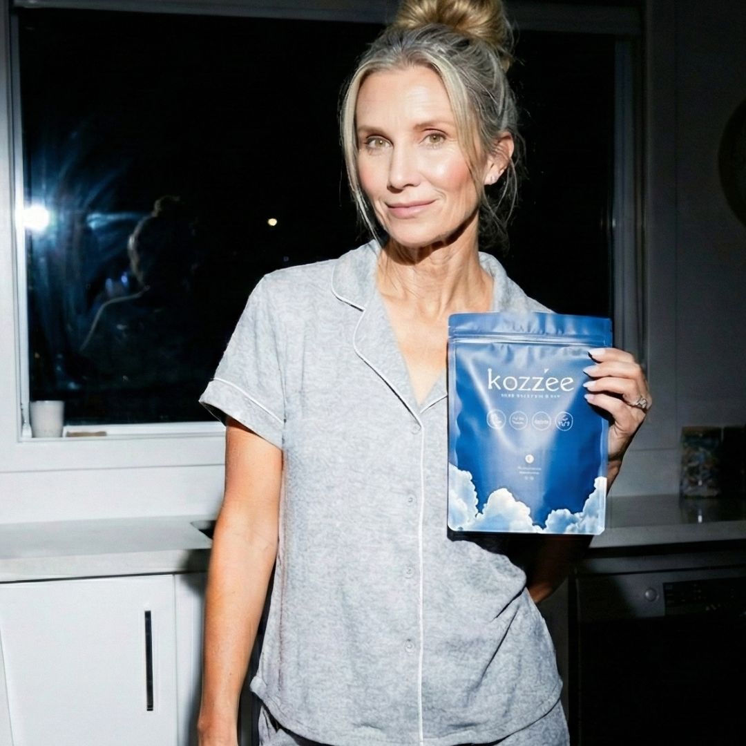 A woman in grey pajamas holds a blue pouch labeled 'kozzēe' while standing in a kitchen at night.