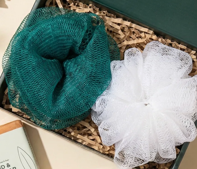 A green and a white mesh bath sponge sit inside a gift box on shredded paper.
