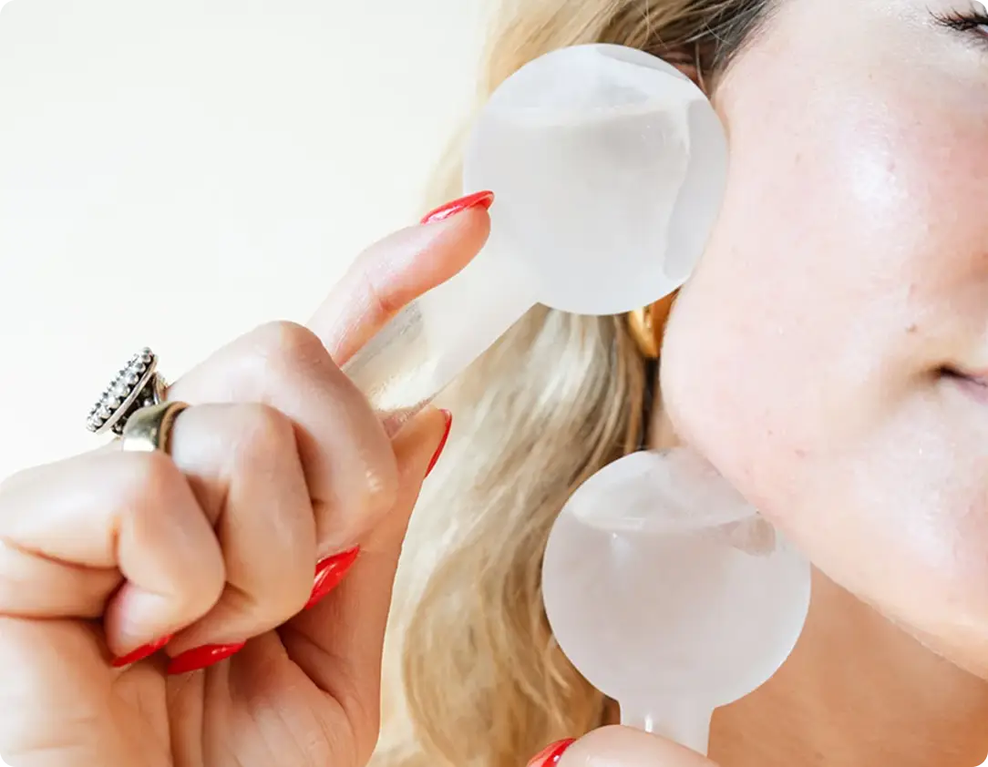 A close-up of a woman with red nails holding two clear ice globes against her cheek.