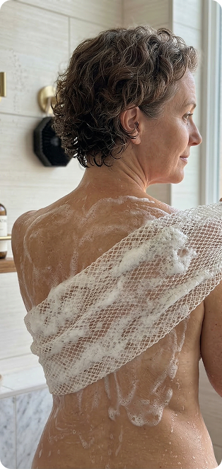 A woman from behind using a soapy, mesh back scrubber in a tiled shower.