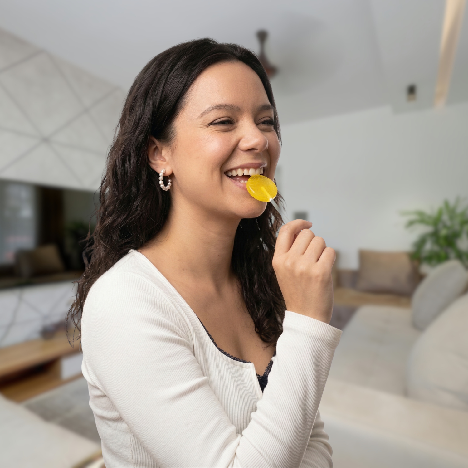 A smiling young woman with long dark hair holds a yellow lollipop in her mouth.