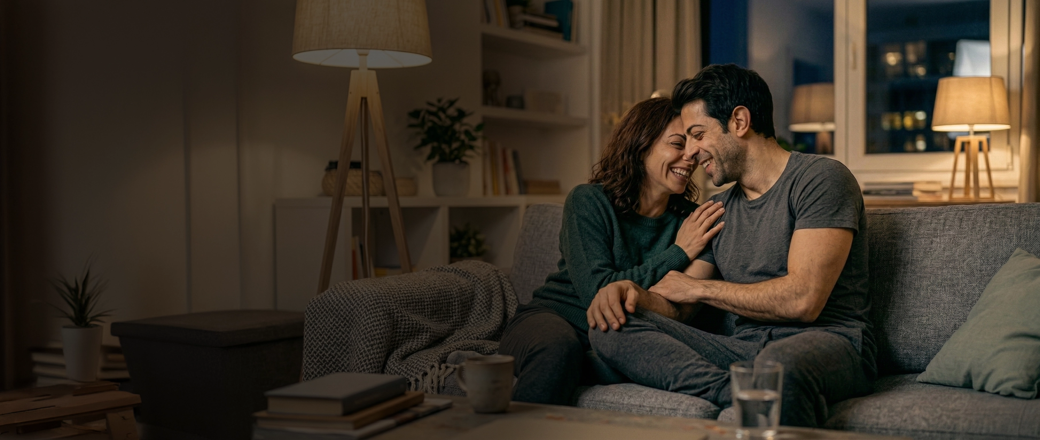 A smiling couple cuddling on a couch in a warmly lit living room at night.