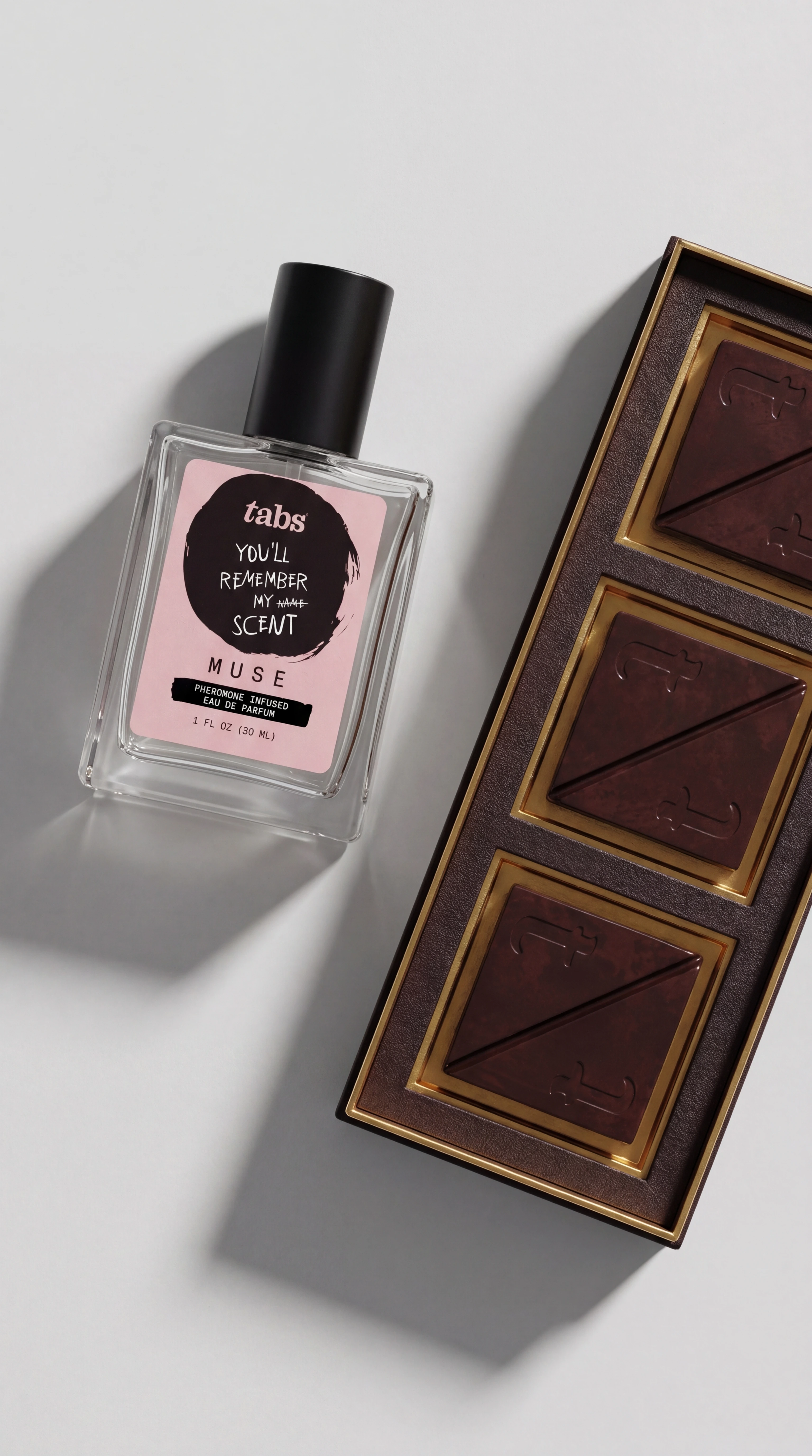 A glass bottle of perfume with a pink label lies next to a box of three chocolate squares.