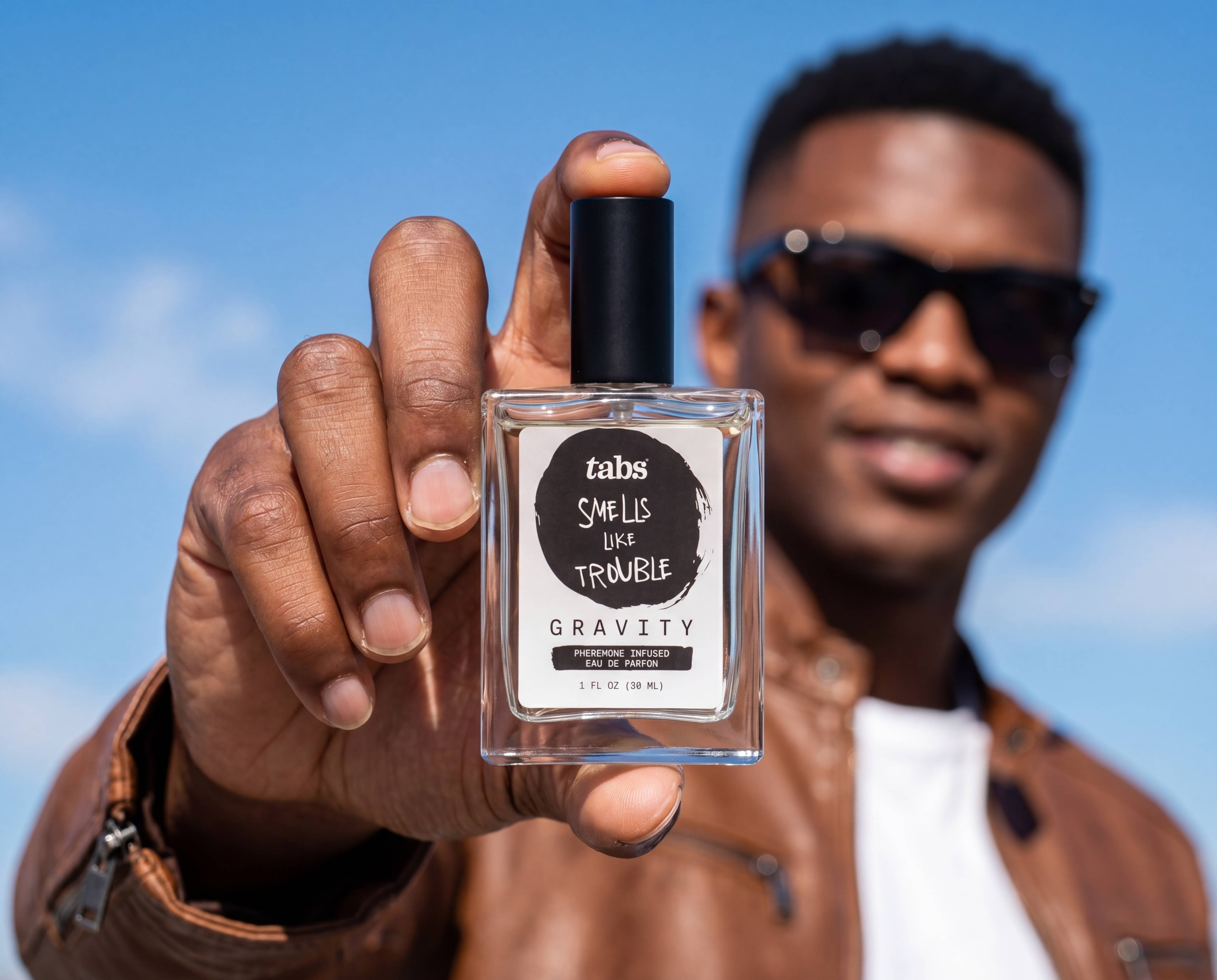 A man holds up a bottle of Tabs Gravity perfume against a blue sky.
