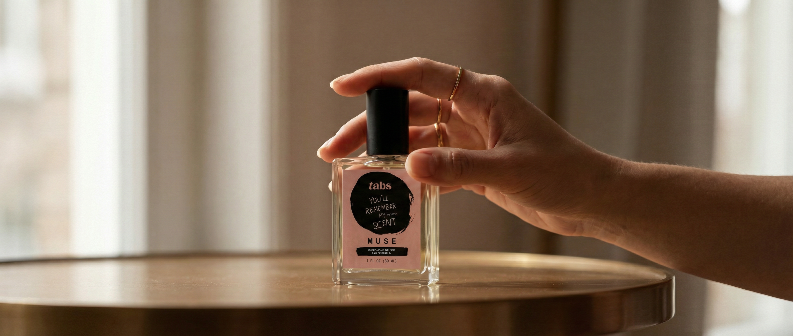 A hand with gold rings holds a rectangular glass bottle of perfume on a wooden table.