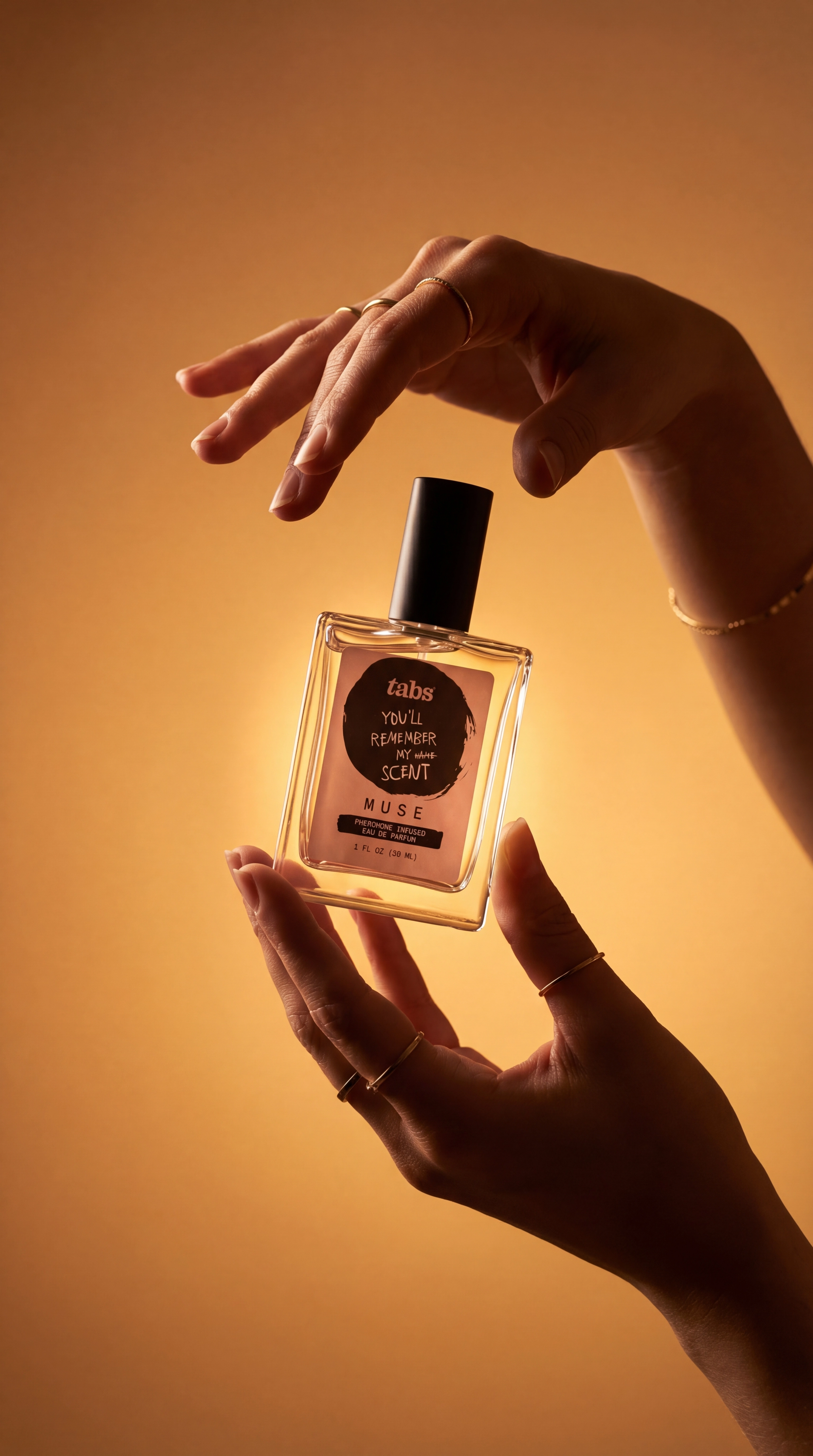 A pair of hands with gold rings holds a bottle of perfume against a warm, golden background.