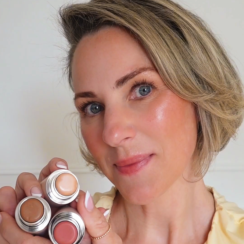A woman with blonde hair and blue eyes holds up three different shades of stick makeup.