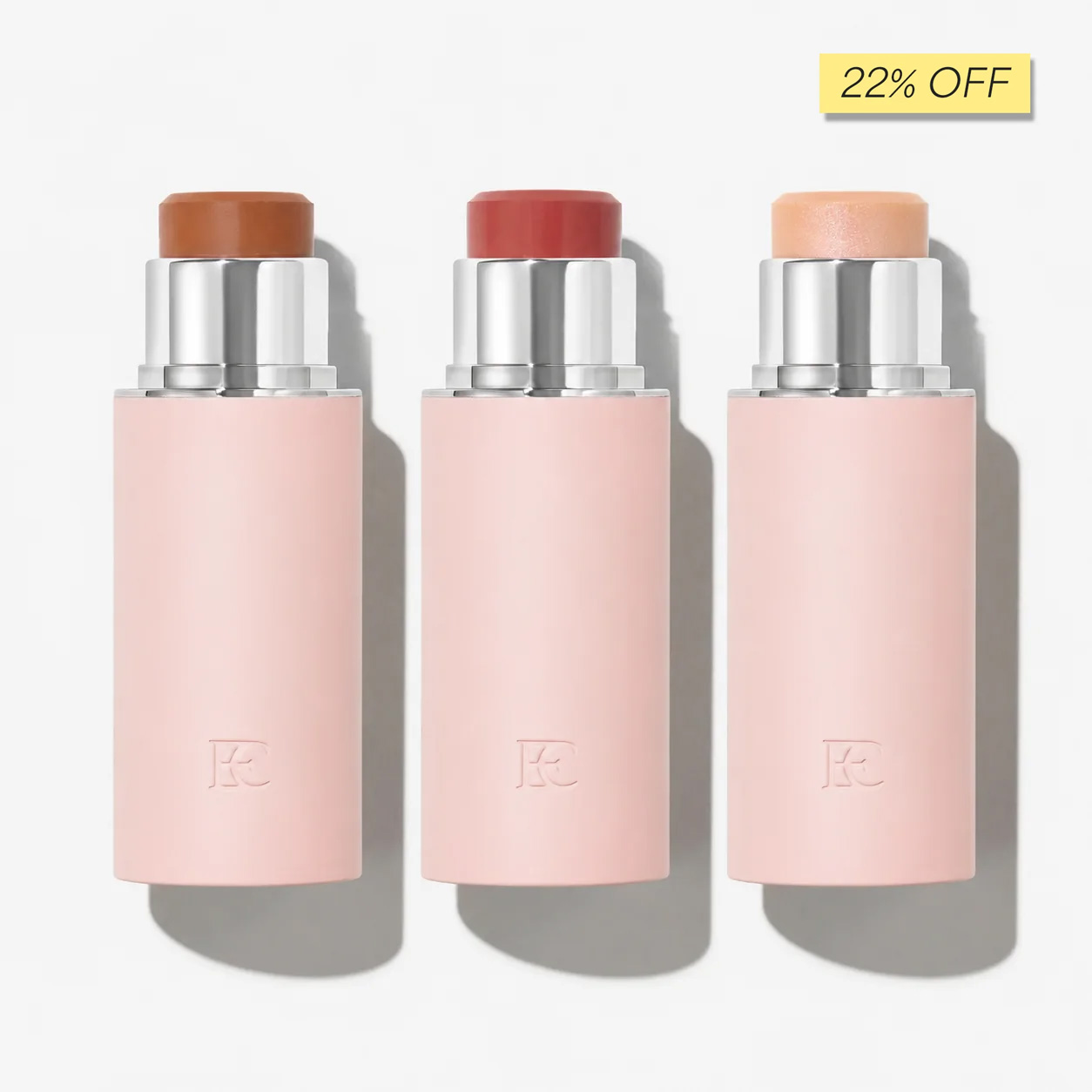 Three makeup sticks in light pink tubes, showing shades of brown, reddish-pink, and light peach.