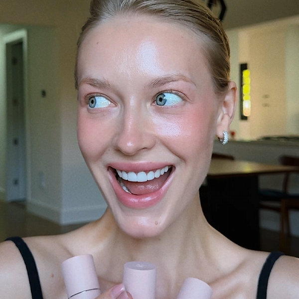 A smiling woman with rosy cheeks holds up three pink makeup sticks.