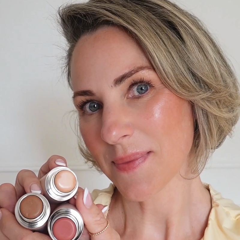 A woman with blonde hair holds up three different makeup sticks to the camera.
