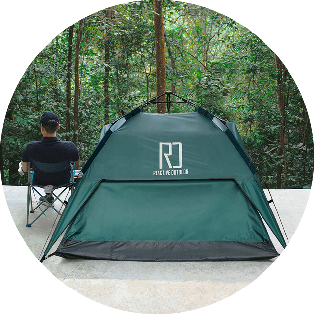 Person sitting next to a green tent in a forest setting.