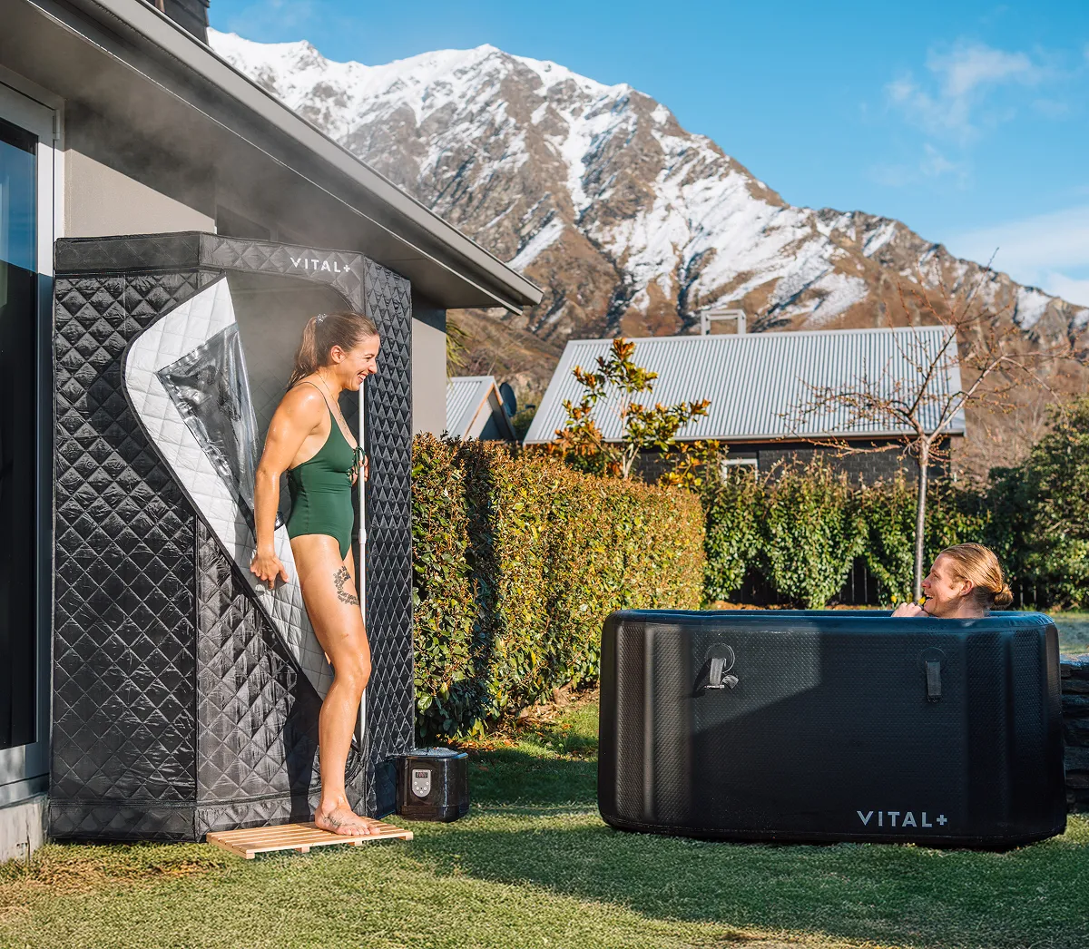 Two women use a portable sauna and an inflatable cold plunge tub outdoors with mountains in the background.