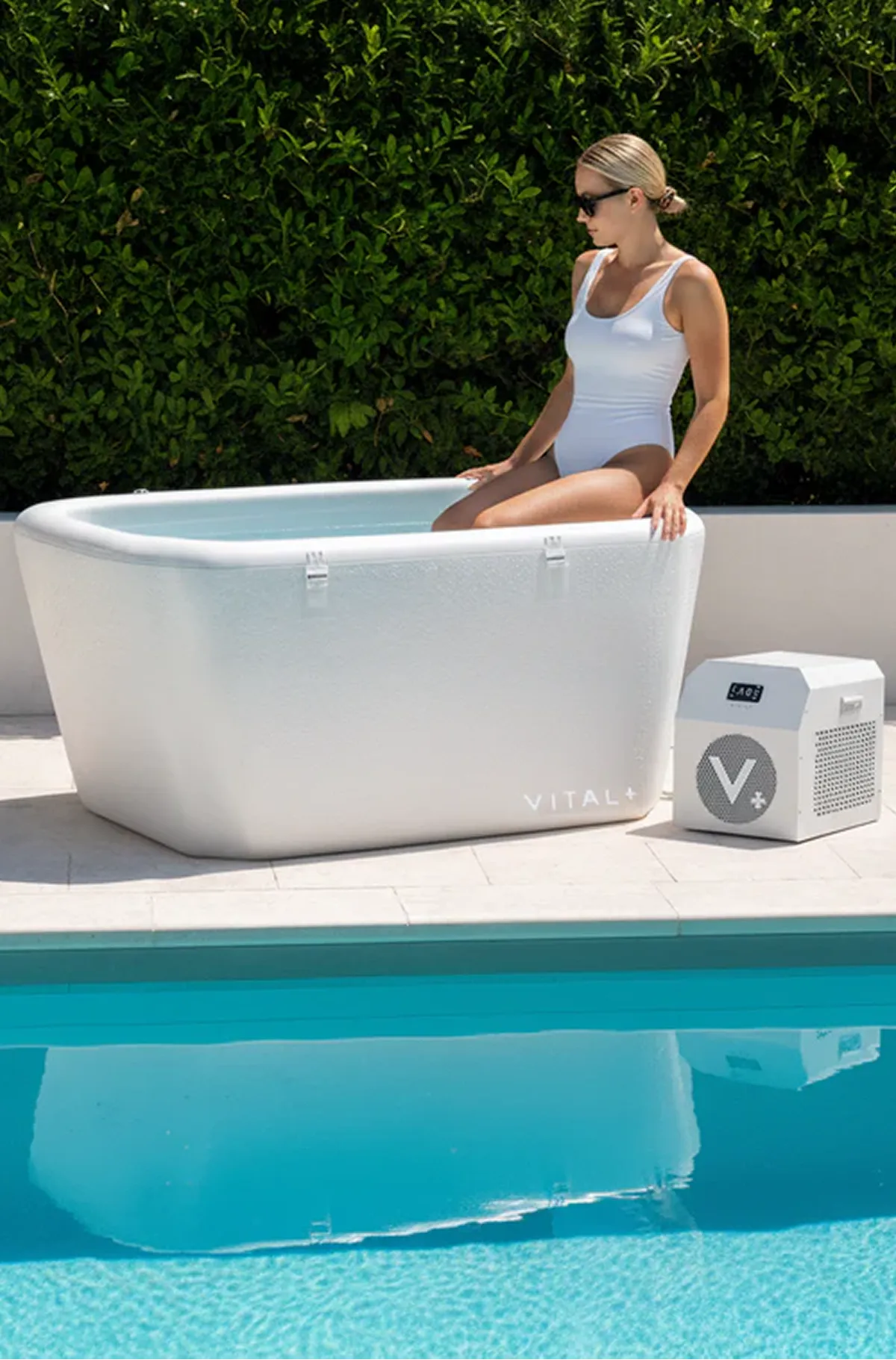 A woman in a white swimsuit sits on the edge of a white portable ice bath by a pool.