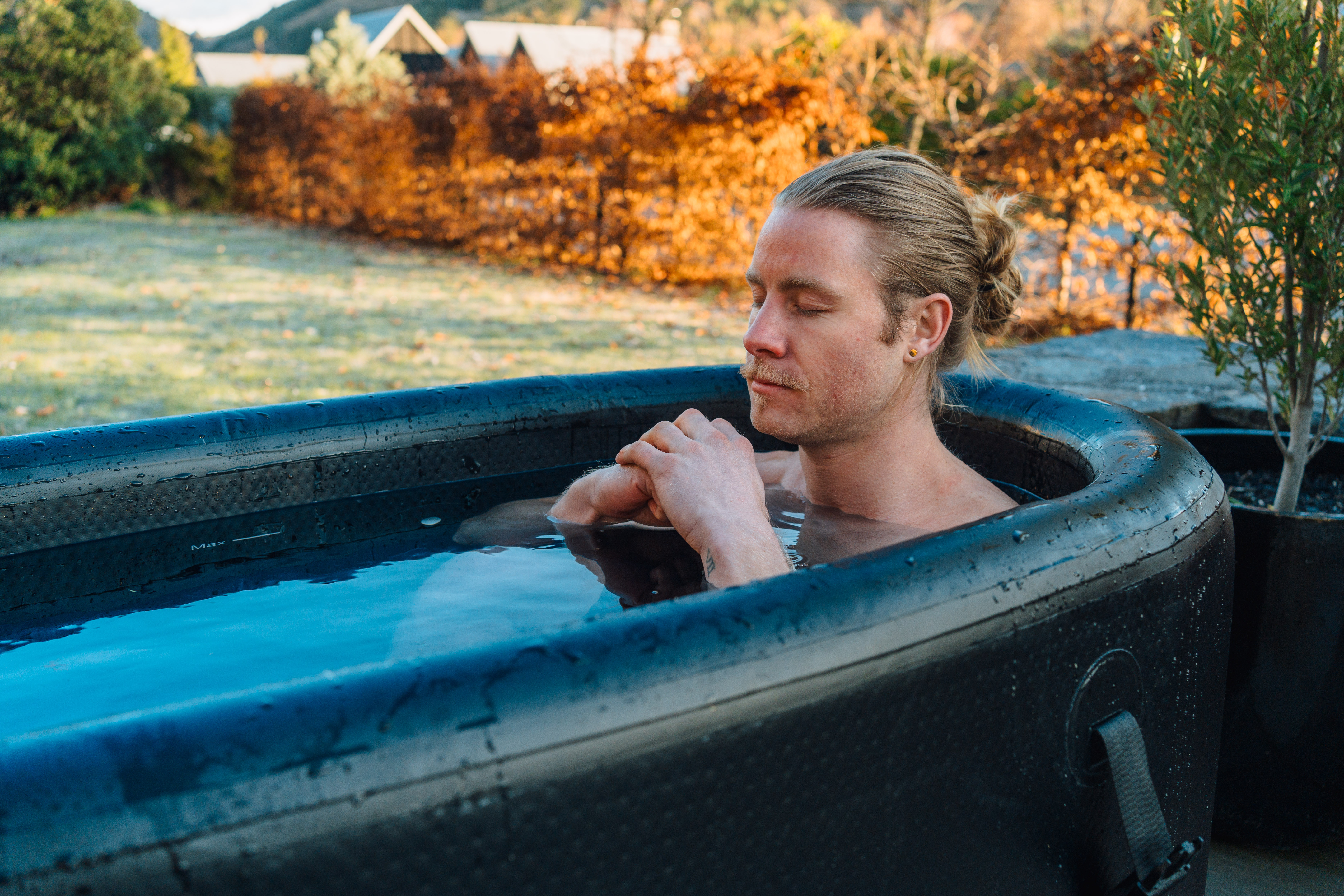 A man with his hair tied back sits in a black inflatable tub outdoors with his eyes closed.