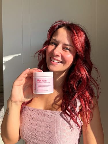 A smiling woman with long red hair holds up a small pink jar of Menora supplements.