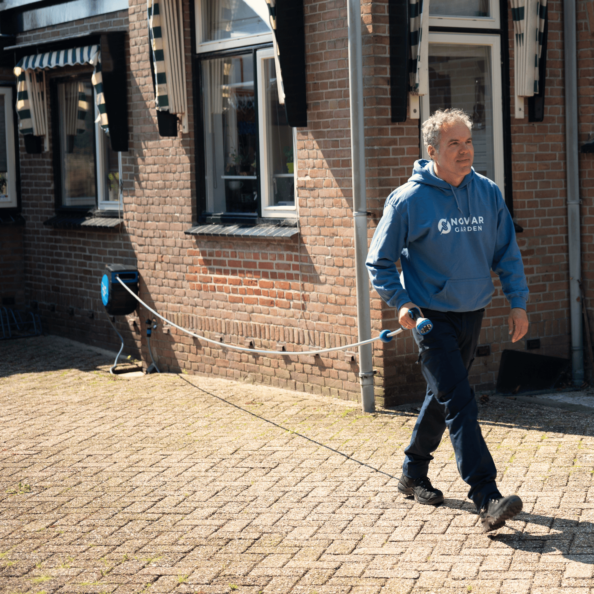Man in blue hoodie walking outside a brick building with a hose reel.
