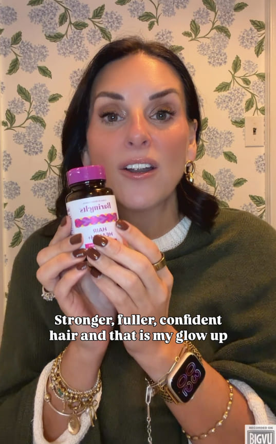 A woman with dark hair holds up a small bottle of supplements in front of a floral wall.