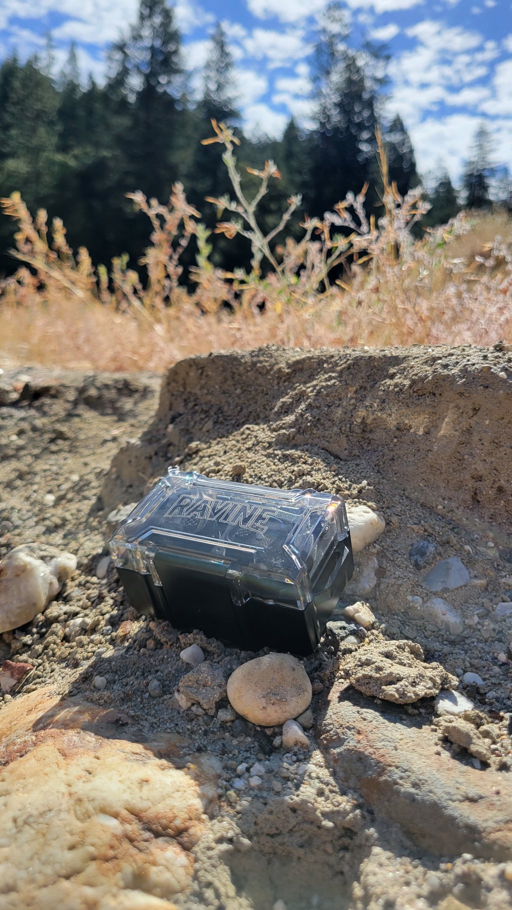 A small black case with a clear lid labeled 'RAVINE' sits on rocky ground outdoors.