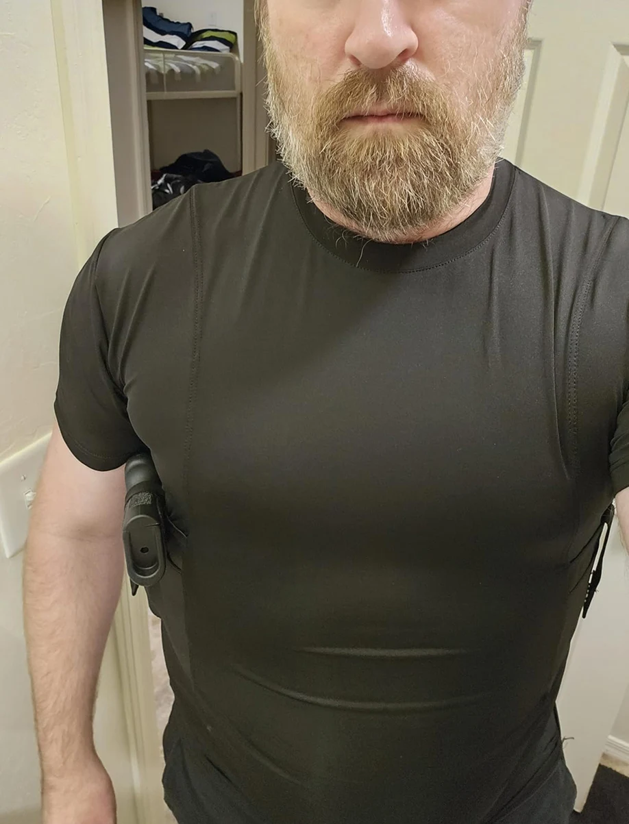 A man with a beard wearing a black concealed carry compression shirt with a handgun holstered under his arm.