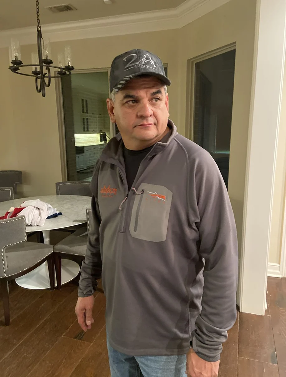 A man in a grey pullover and a baseball cap stands indoors in a dining area, looking off-camera.