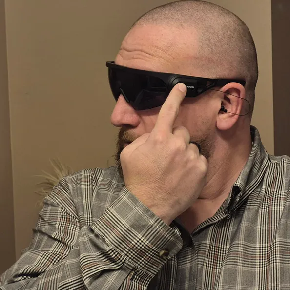 A bald man with a beard wearing a plaid shirt and smart sunglasses with an attached earbud.