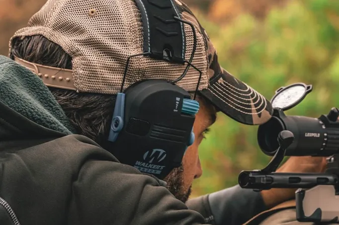 Person in cap wearing earmuffs aiming a rifle outdoors.