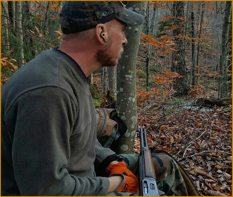 Person sitting in a forest holding a rifle, wearing a cap and orange gloves.