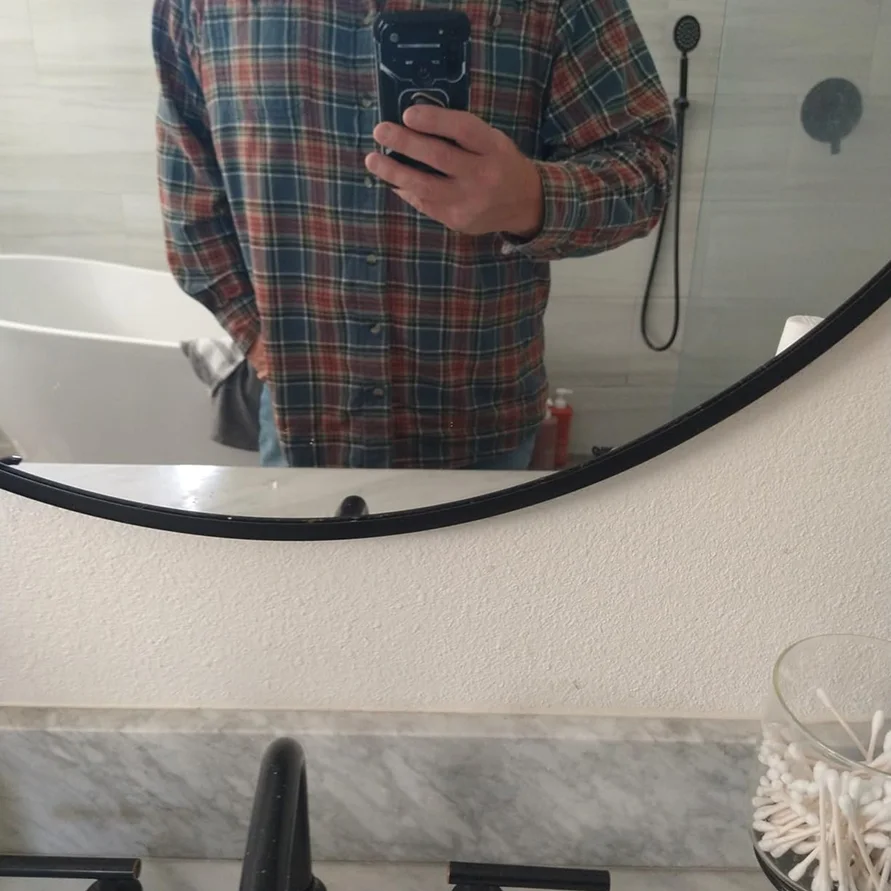 A person in a plaid shirt takes a selfie in a bathroom mirror, showing a bathtub in the reflection.