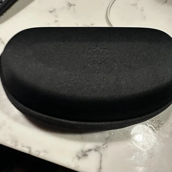 A black, fabric-covered, hard-shell glasses case with an embossed logo sits on a marble countertop.