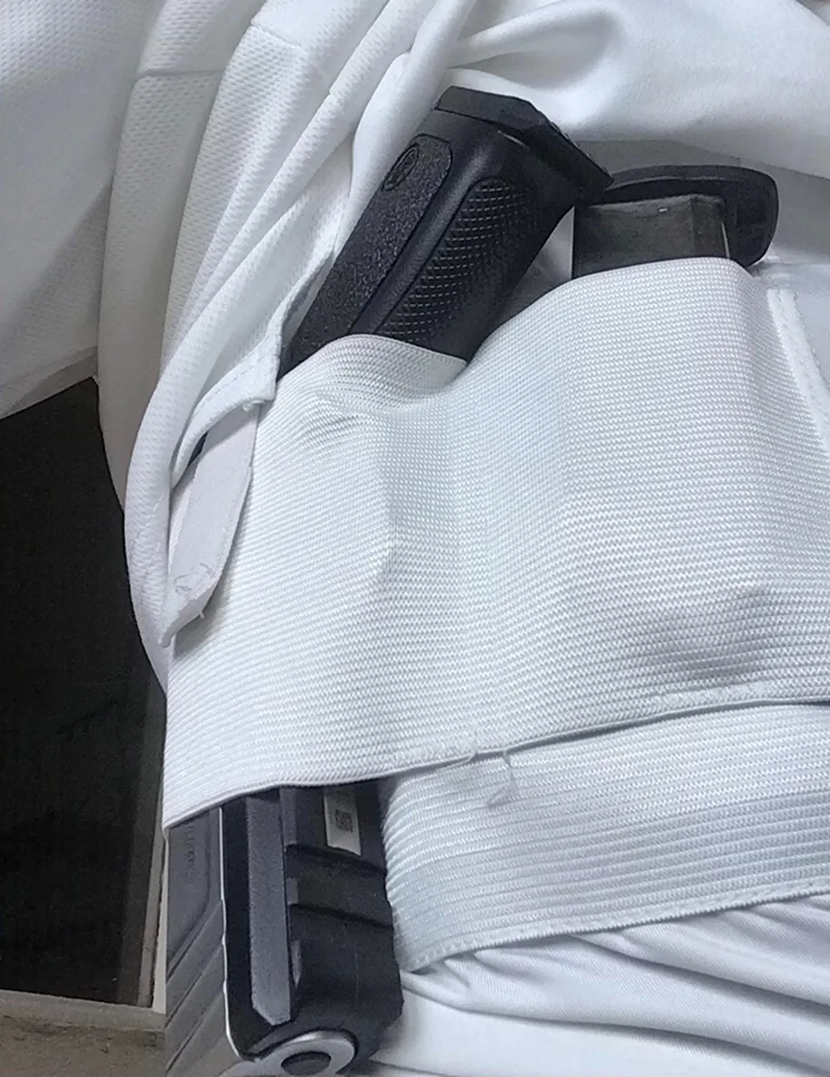 A close-up of a black handgun tucked into a white elastic belly band holster under a white shirt.