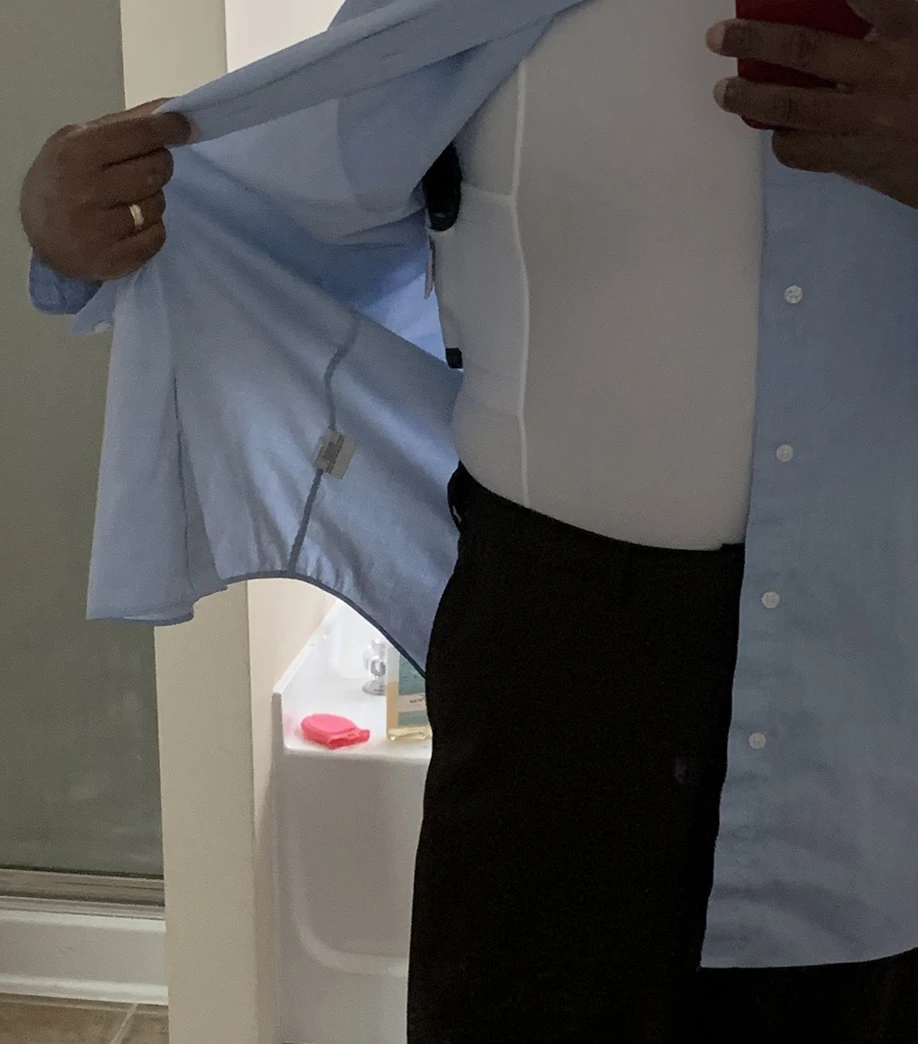 A person lifts their blue shirt to reveal a handgun in a white shoulder holster undershirt.