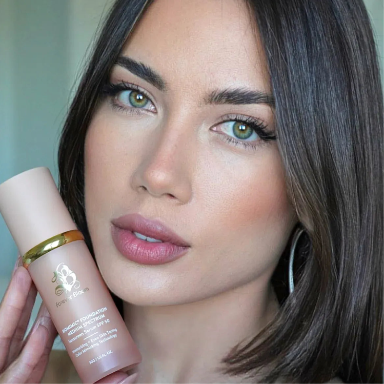 A woman with green eyes and brown hair, wearing full makeup, holds up a pink bottle of foundation.