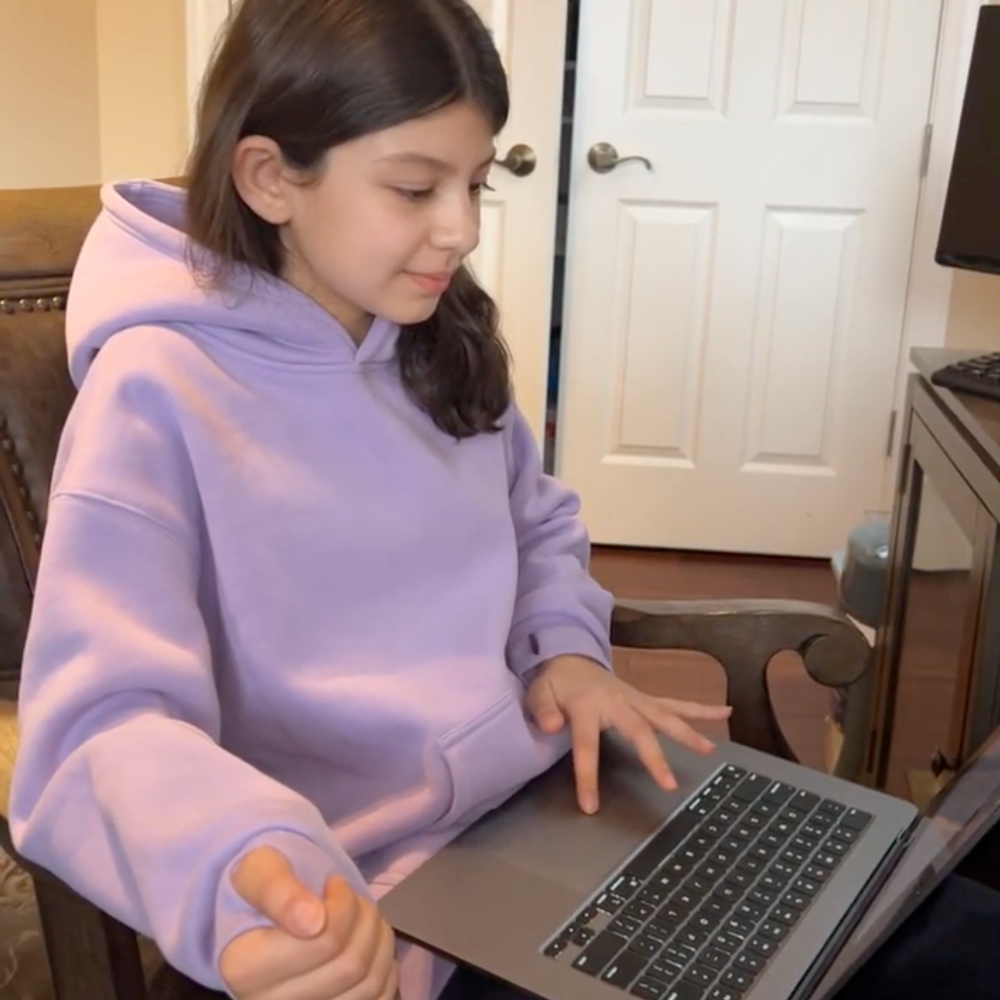 Young girl in a purple hoodie using a laptop at a desk.