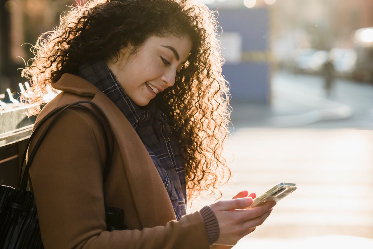Woman with curly hair smiling at her phone outdoors.