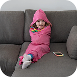 A young child wrapped tightly in a pink hooded blanket sits on a grey couch next to a smartphone.