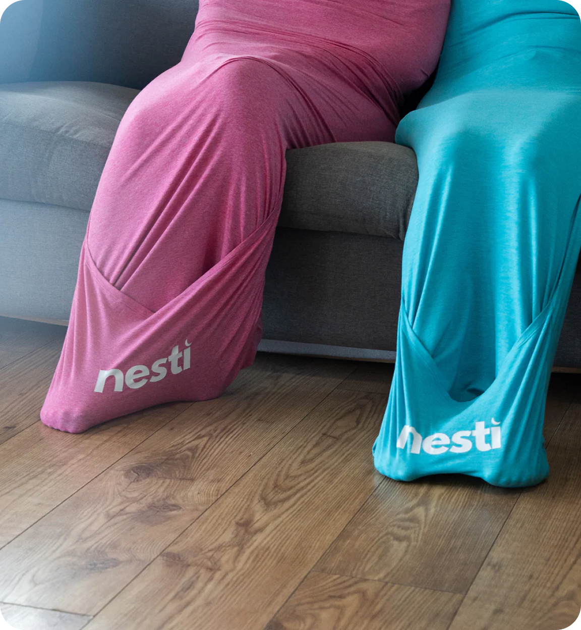 Two people sit on a couch fully enclosed in pink and blue stretchy fabric sacks with 'nesti' logos.
