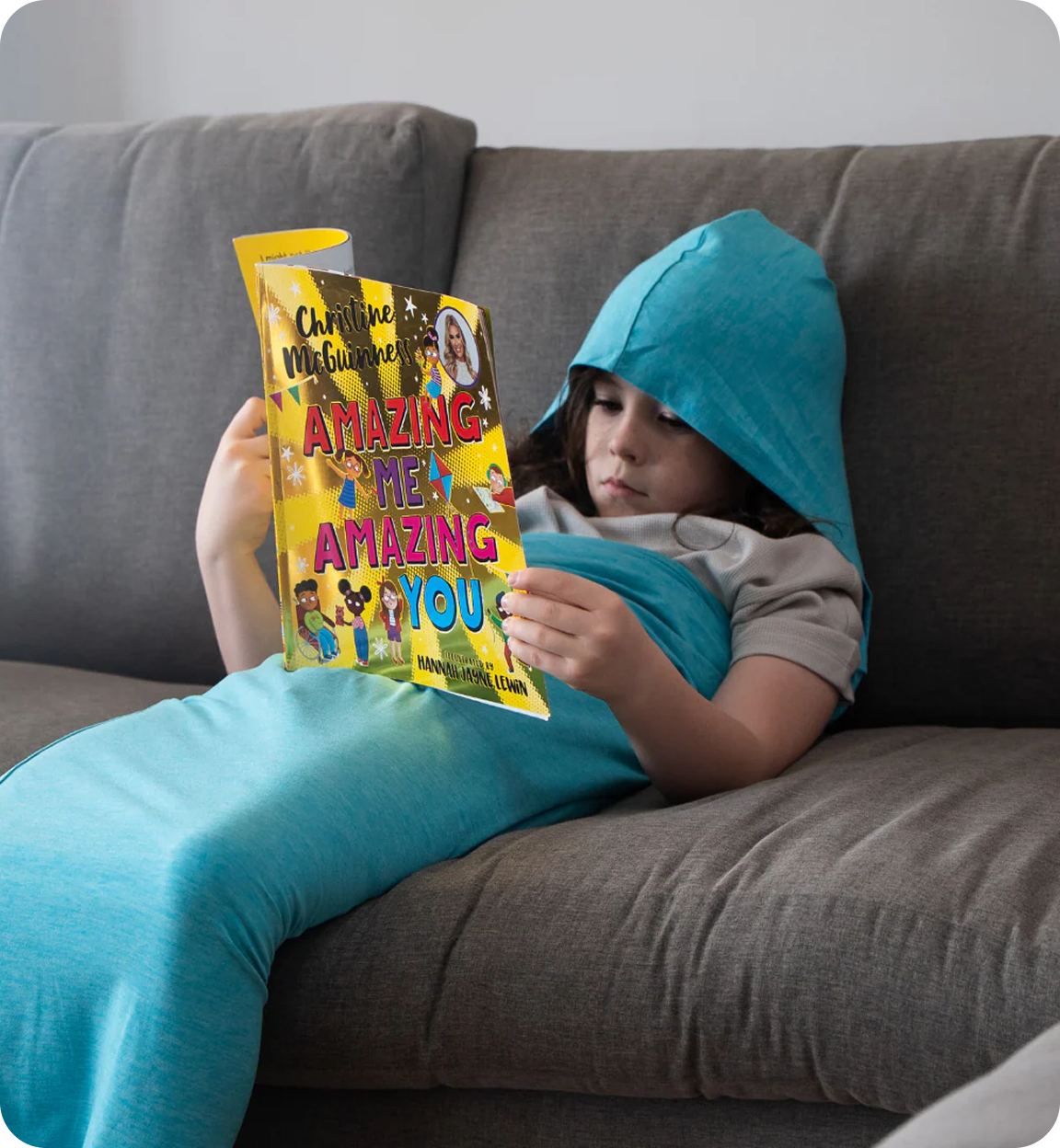 A child wearing a blue hooded blanket lies on a couch and reads a book.