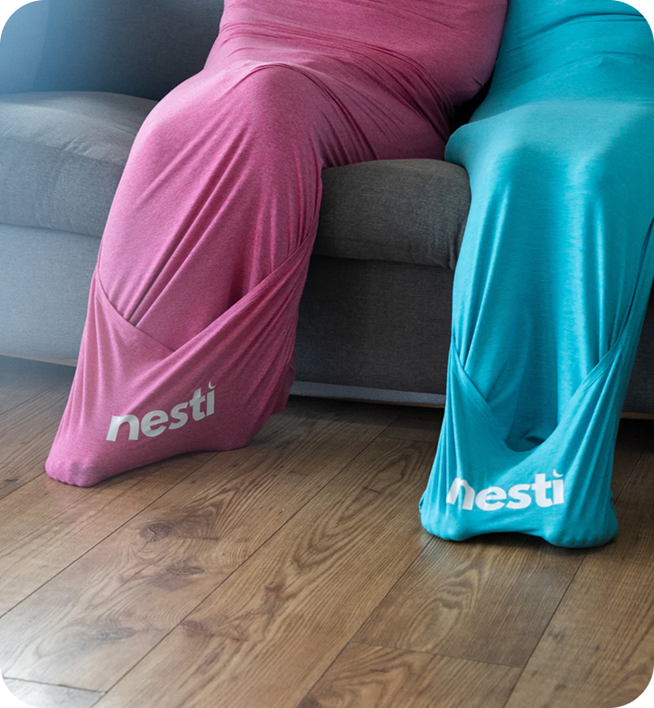Two people sit on a couch, each enclosed in a large, stretchy fabric sack, one pink, one turquoise.