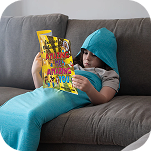 A child in a blue hooded mermaid tail blanket reads a colorful book on a gray couch.