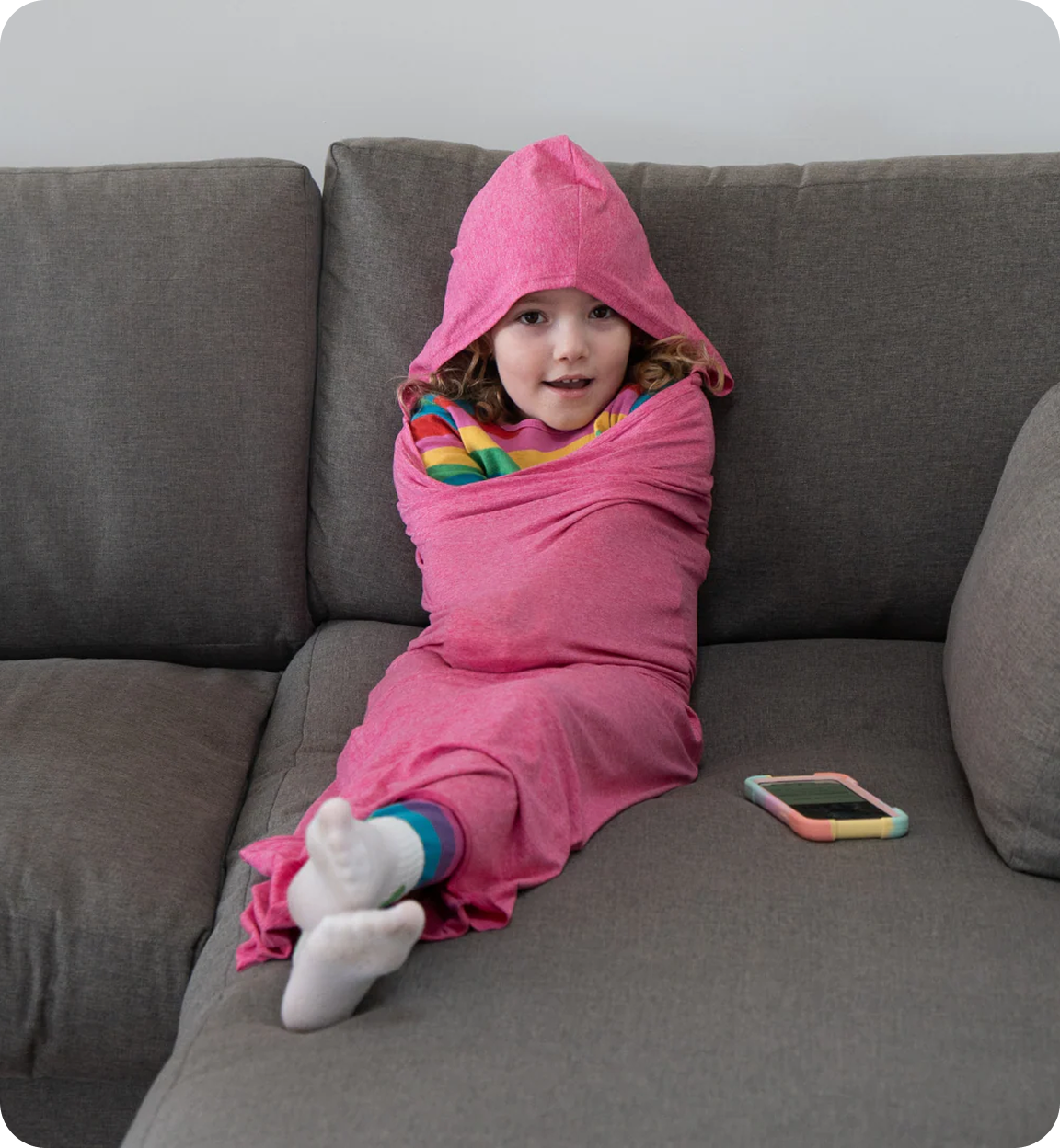 A young girl wrapped snugly in a pink hooded blanket sits on a grey couch looking at the camera.