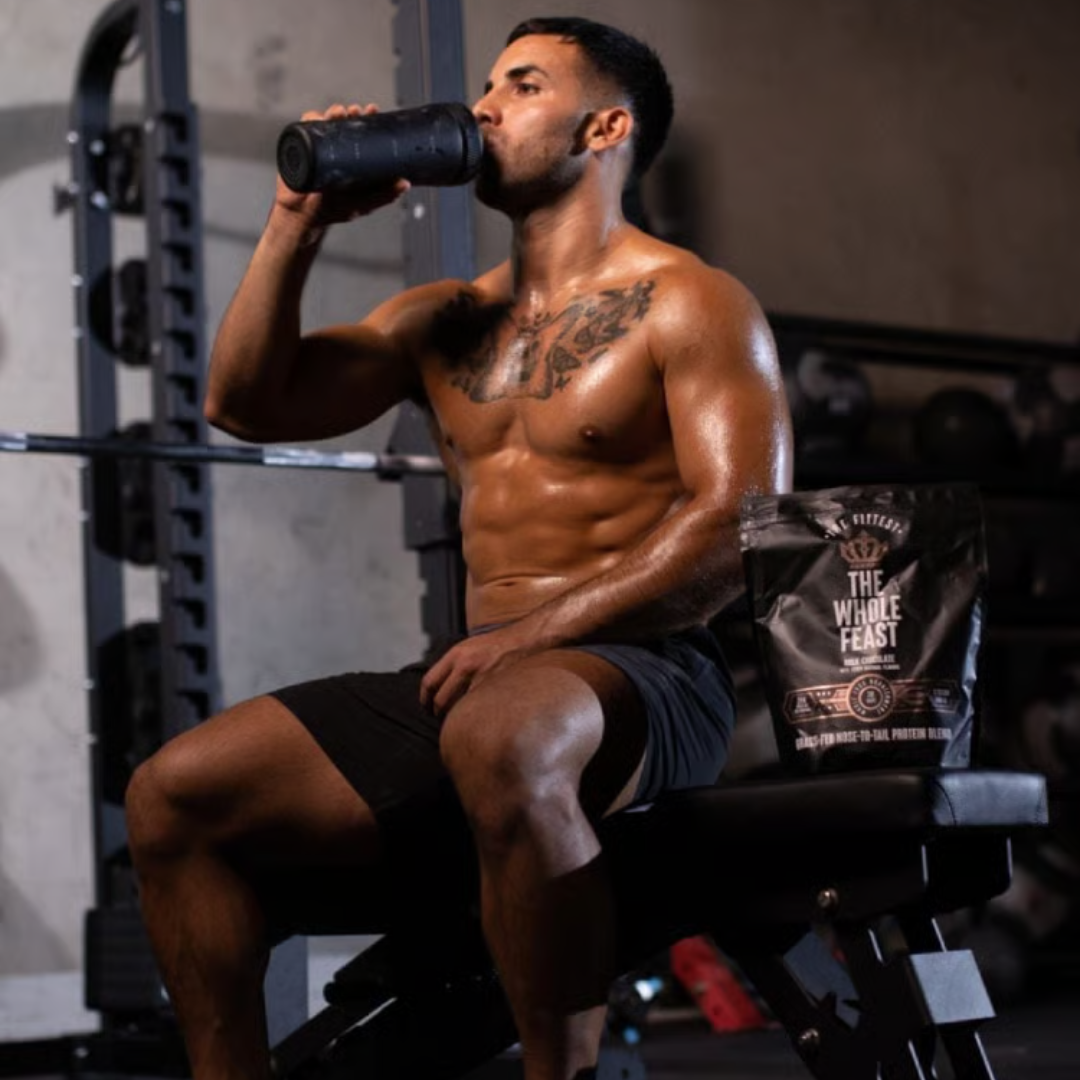 A muscular man with tattoos sits shirtless on a gym bench, drinking from a black shaker bottle.
