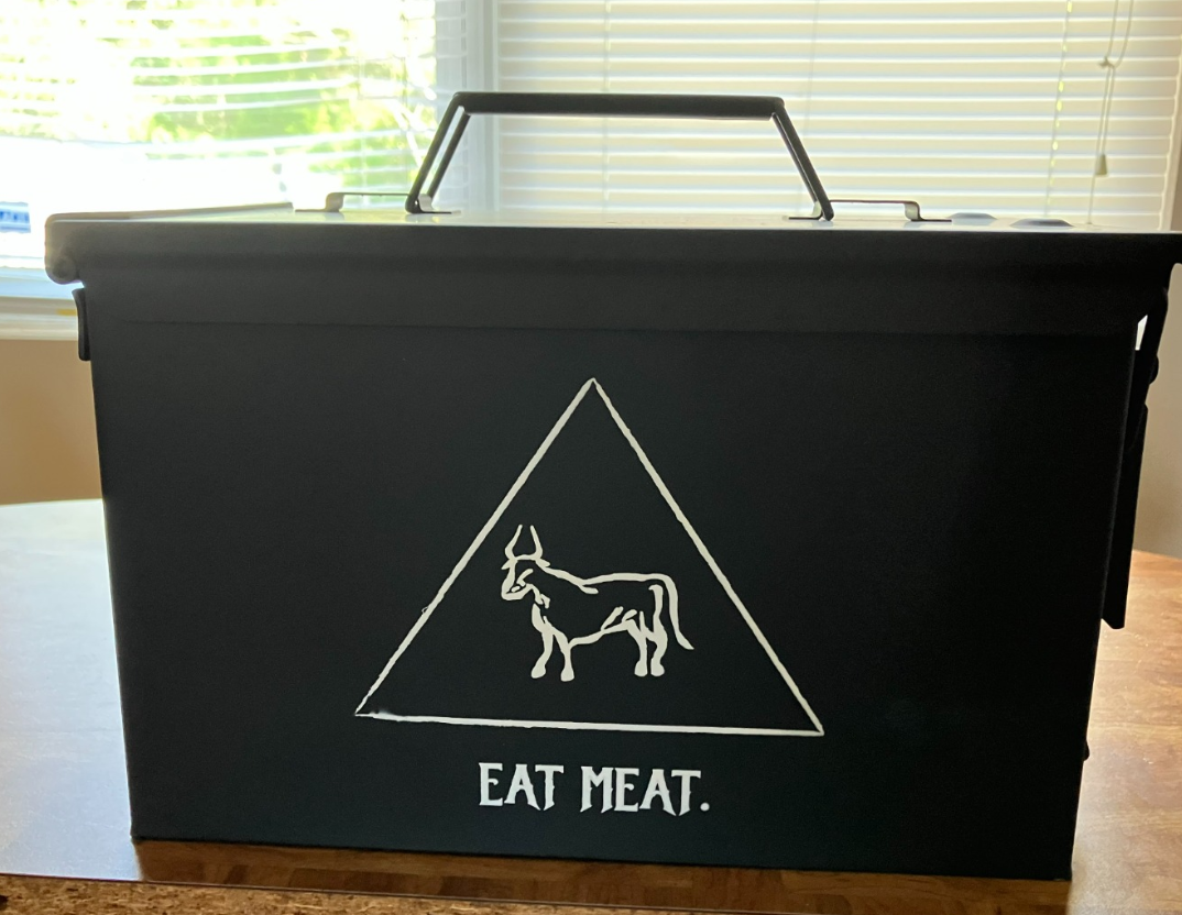 A black metal ammo can with a white graphic of a bull in a triangle and the words 'EAT MEAT.'