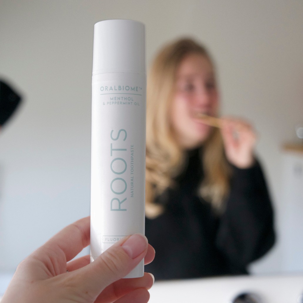 A hand holds a tube of toothpaste, with a blurred person brushing their teeth in the background.