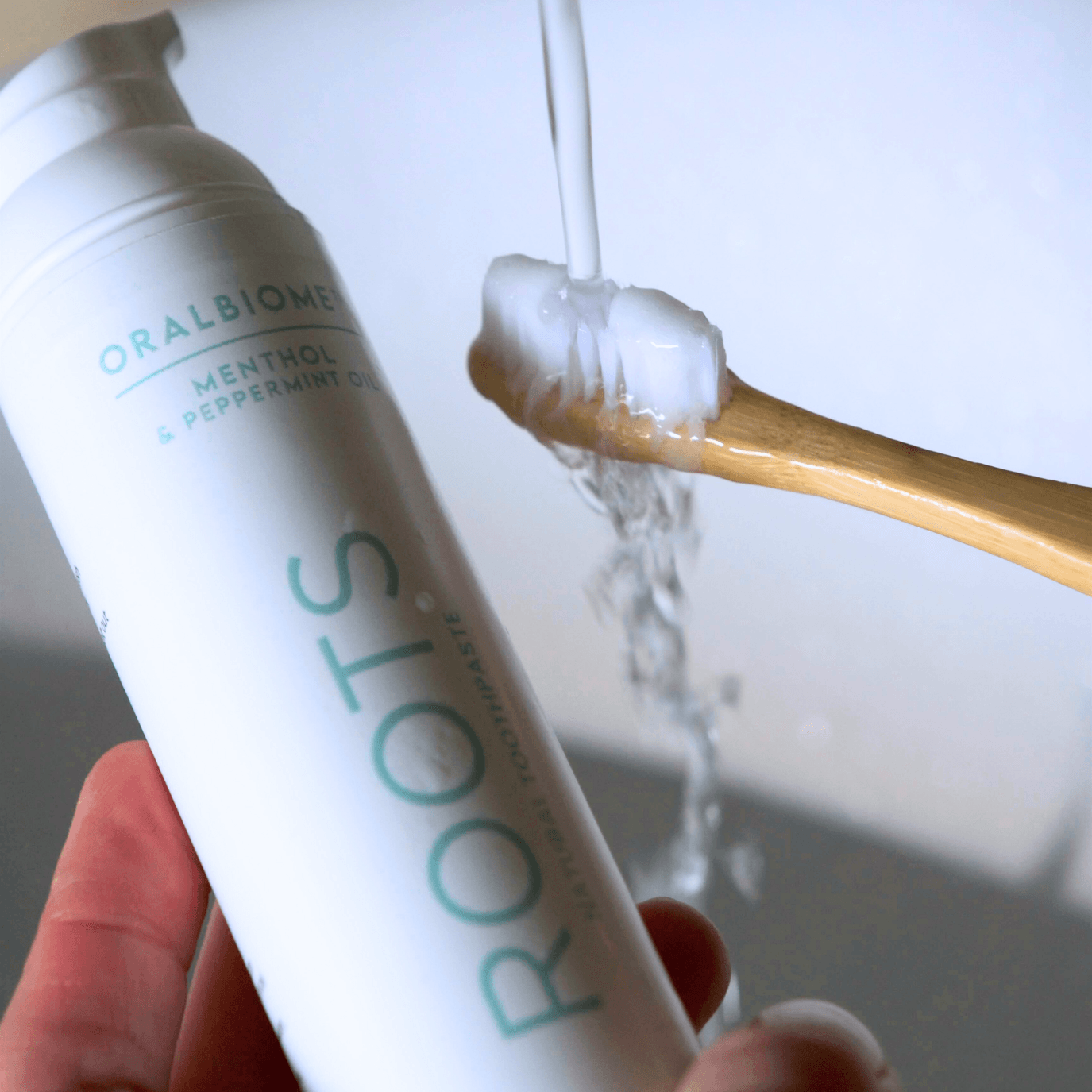 A hand holds a bottle of Oralbiome toothpaste next to a bamboo toothbrush being rinsed under water.