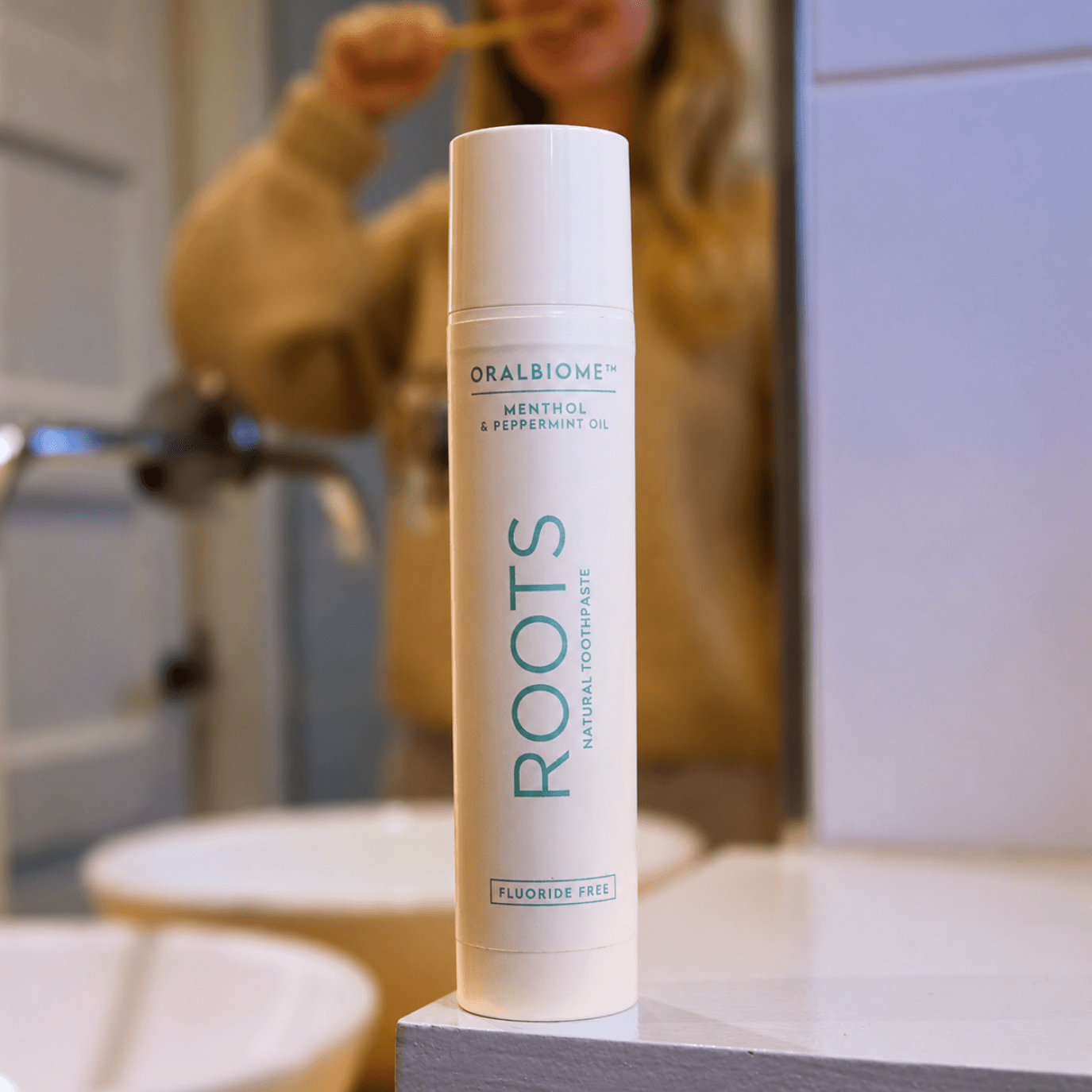 A bottle of Roots toothpaste on a bathroom counter, with a person brushing their teeth in the background.