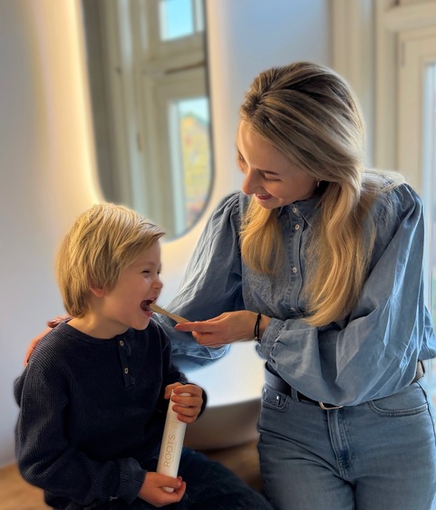 A woman helps a young, blond-haired boy brush his teeth in a bathroom.