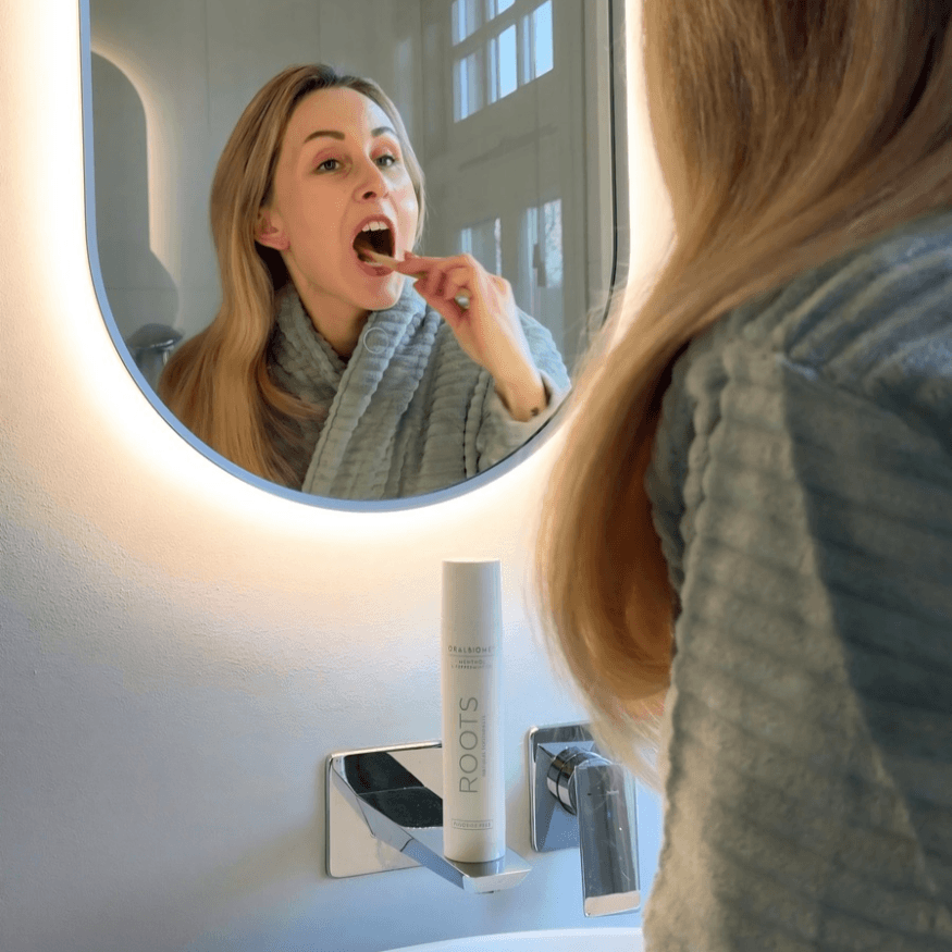 A woman in a bathrobe brushes her teeth while looking into an illuminated bathroom mirror.