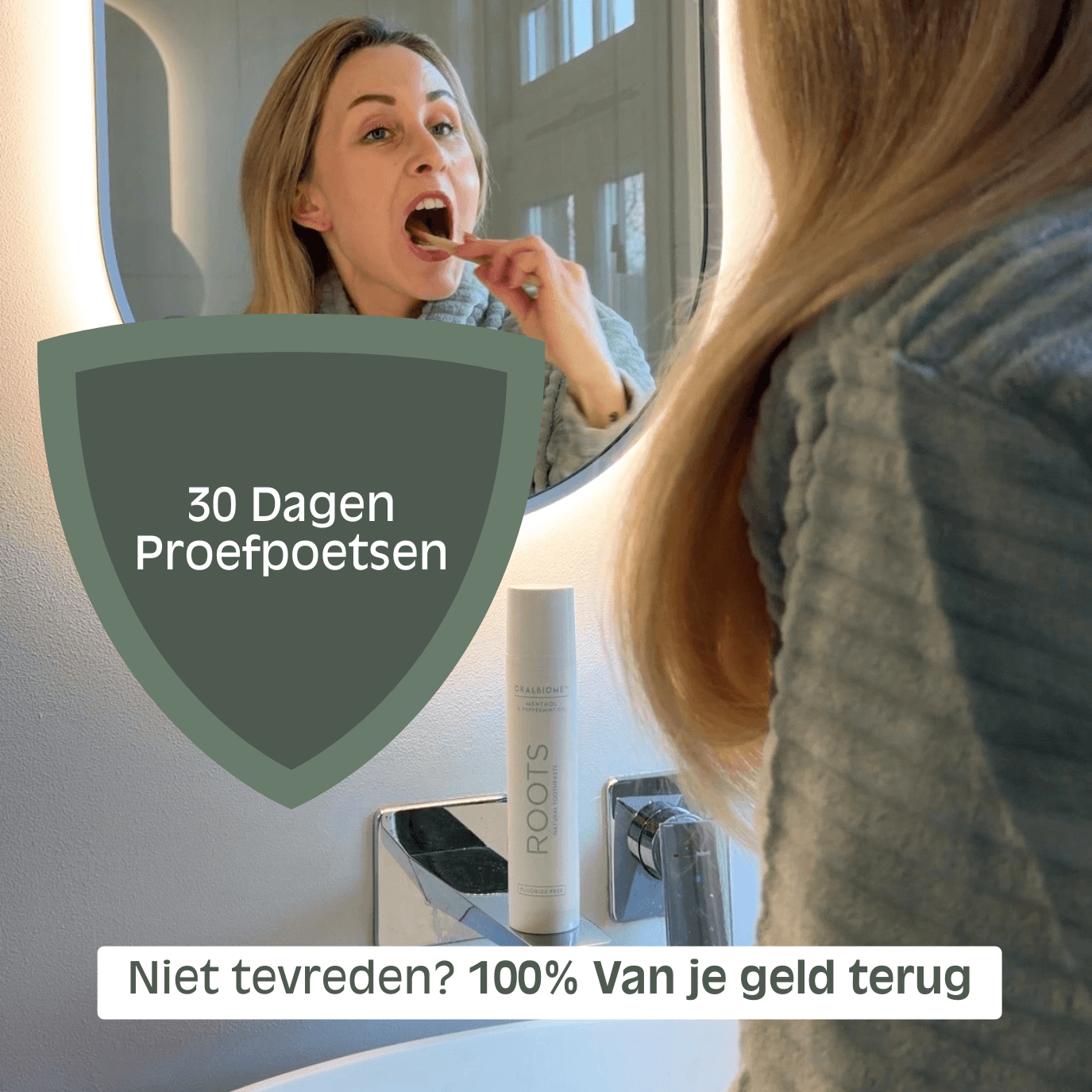 A woman brushing her teeth while looking at her reflection in a bathroom mirror next to a tube of toothpaste.
