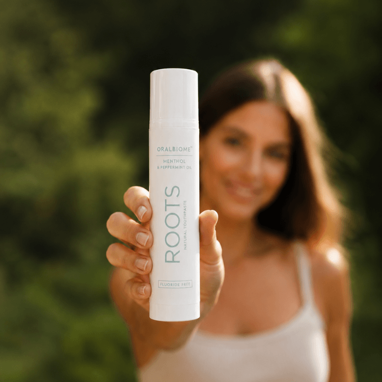 A woman holds a white tube of Oralbiome Roots toothpaste towards the camera, with a blurred green background.